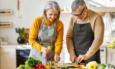 Casal de adultos na cozinha, preparando uma salada com pepino, alface, rabanete e outros vegetais.