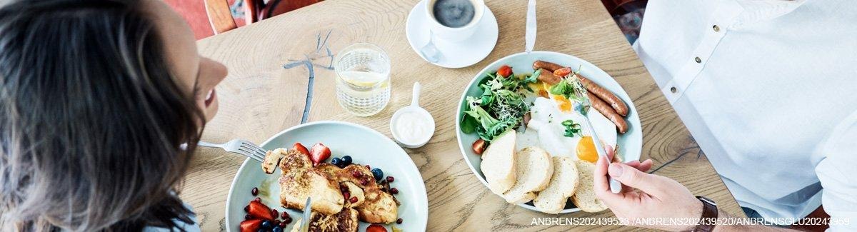 Casal de adultos comendo um café da manhã saudável de: ovos, salsicha, pão, morangos, mirtilos, torradas francesas e mais.