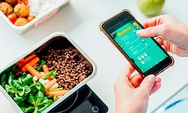 Mulher utilizando um aplicativo de registro de refeições com um almoço à sua frente em uma mesa.