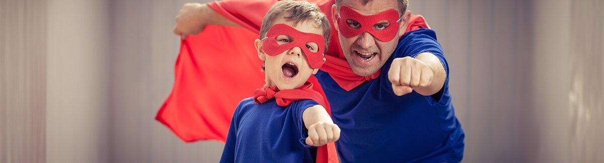 Duas pessoas, um adulto e uma criança, vestindo camisetas azuis e capas vermelhas, posando como se fossem super-heróis com os punhos estendidos para a frente