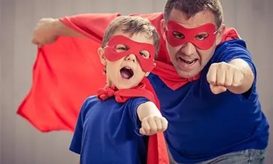 Duas pessoas, um adulto e uma criança, vestindo camisetas azuis e capas vermelhas, posando como se fossem super-heróis com os punhos estendidos para a frente