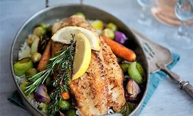 Um prato colorido com um pedaço de peixe grelhado decorado com microvegetais, servido com uma variedade de legumes, incluindo rabanetes, cenouras, ervilhas e batatas, todos arranjados em um prato azul.