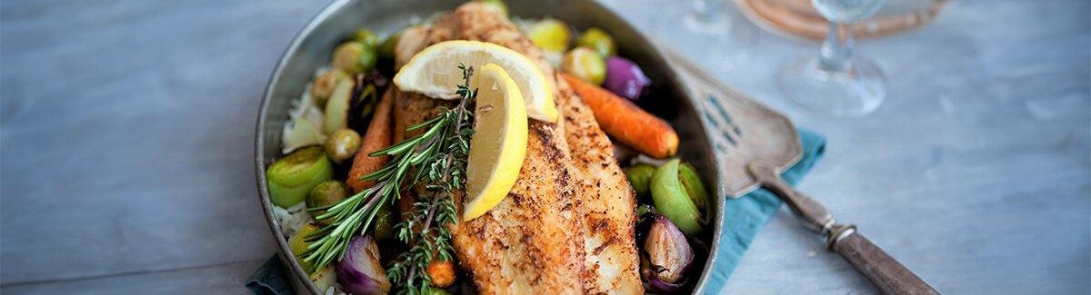 Um prato colorido com um pedaço de peixe grelhado decorado com microvegetais, servido com uma variedade de legumes, incluindo rabanetes, cenouras, ervilhas e batatas, todos arranjados em um prato azul.