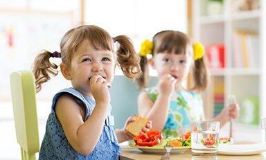 Copo de vitamina de banana com um canudo azul de bolinhas. A vitamina está colocada em uma superfície de madeira, com um cacho de bananas e um pote de mel ao fundo.