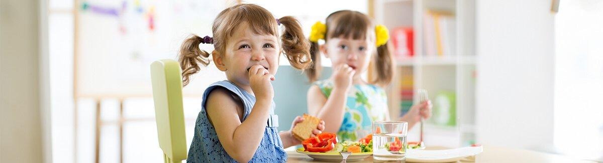 Copo de vitamina de banana com um canudo azul de bolinhas. A vitamina está colocada em uma superfície de madeira, com um cacho de bananas e um pote de mel ao fundo.