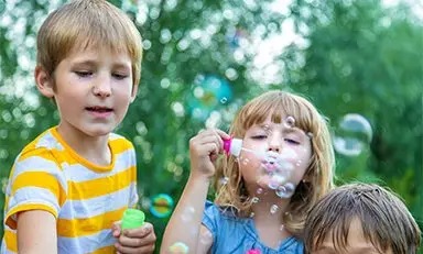 Criança com camiseta laranja brincando com bolhas ao ar livre. Ao fundo, há outras crianças e uma mesa com vários itens, sugerindo uma festa ou reunião. A imagem inclui os logotipos da Abbott e da PediaSure