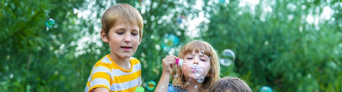 Criança com camiseta laranja brincando com bolhas ao ar livre. Ao fundo, há outras crianças e uma mesa com vários itens, sugerindo uma festa ou reunião. A imagem inclui os logotipos da Abbott e da PediaSure